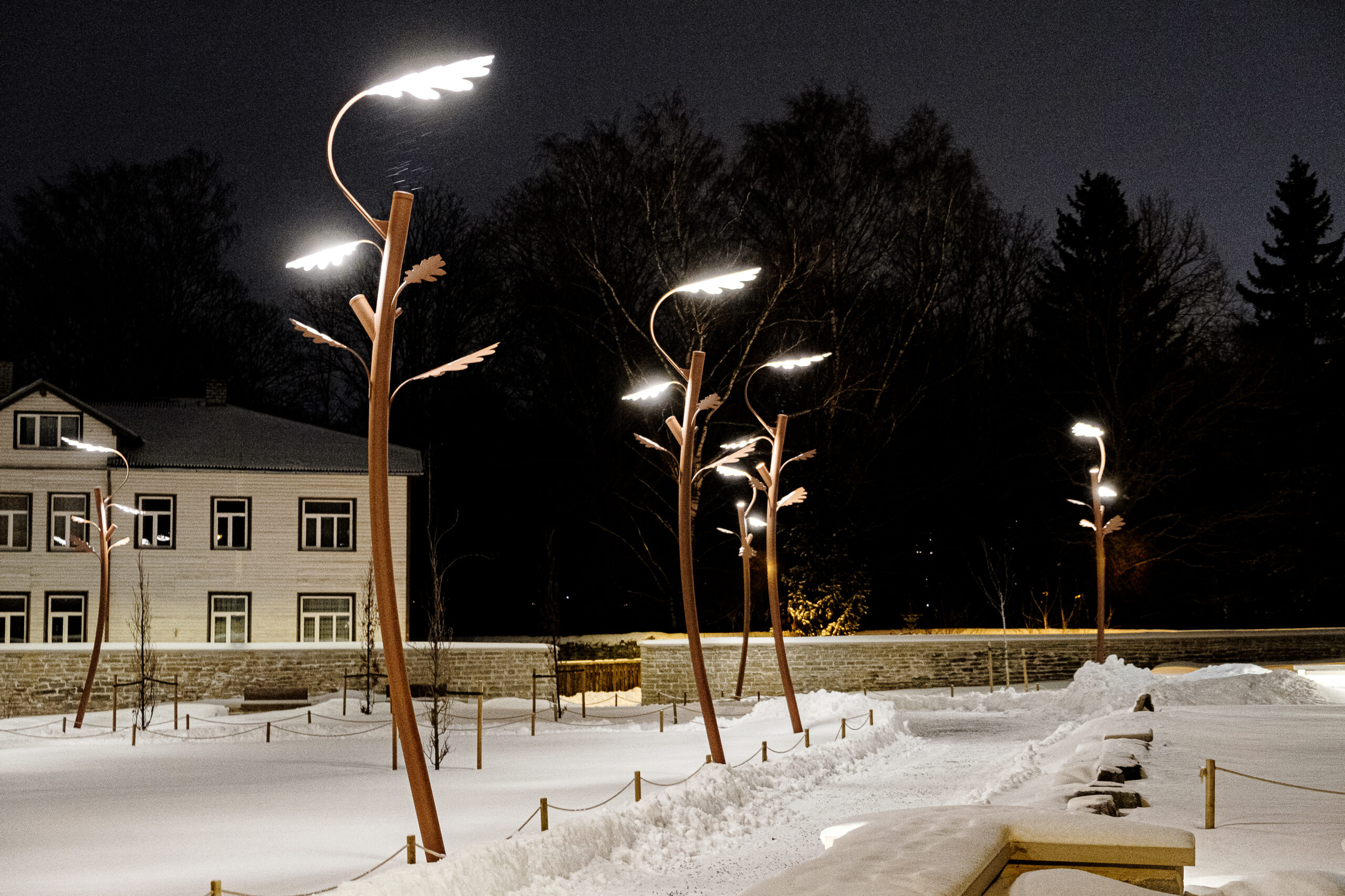 Snow-covered garden at night with illuminated metal oak tree-shaped outdoor pole light, a stone wall, and a house in the background.