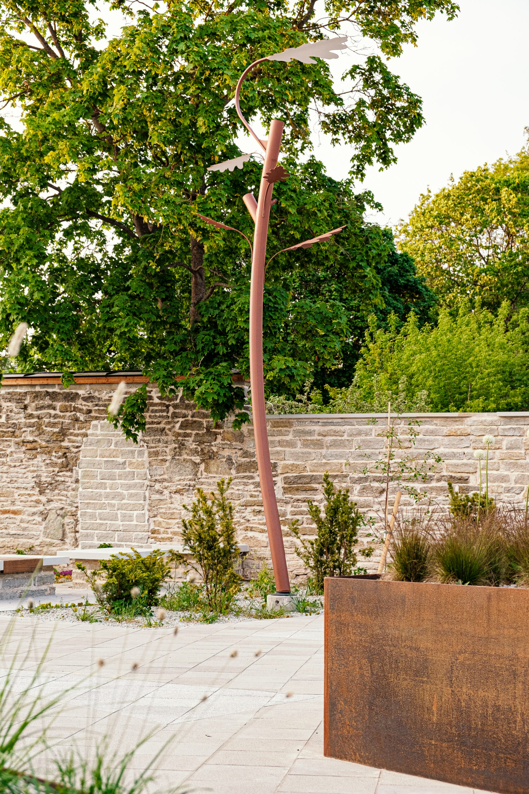 Oak tree shaped outdoor space pole light in a modern garden with a stone wall, greenery, and a large rust-colored planter under a clear sky