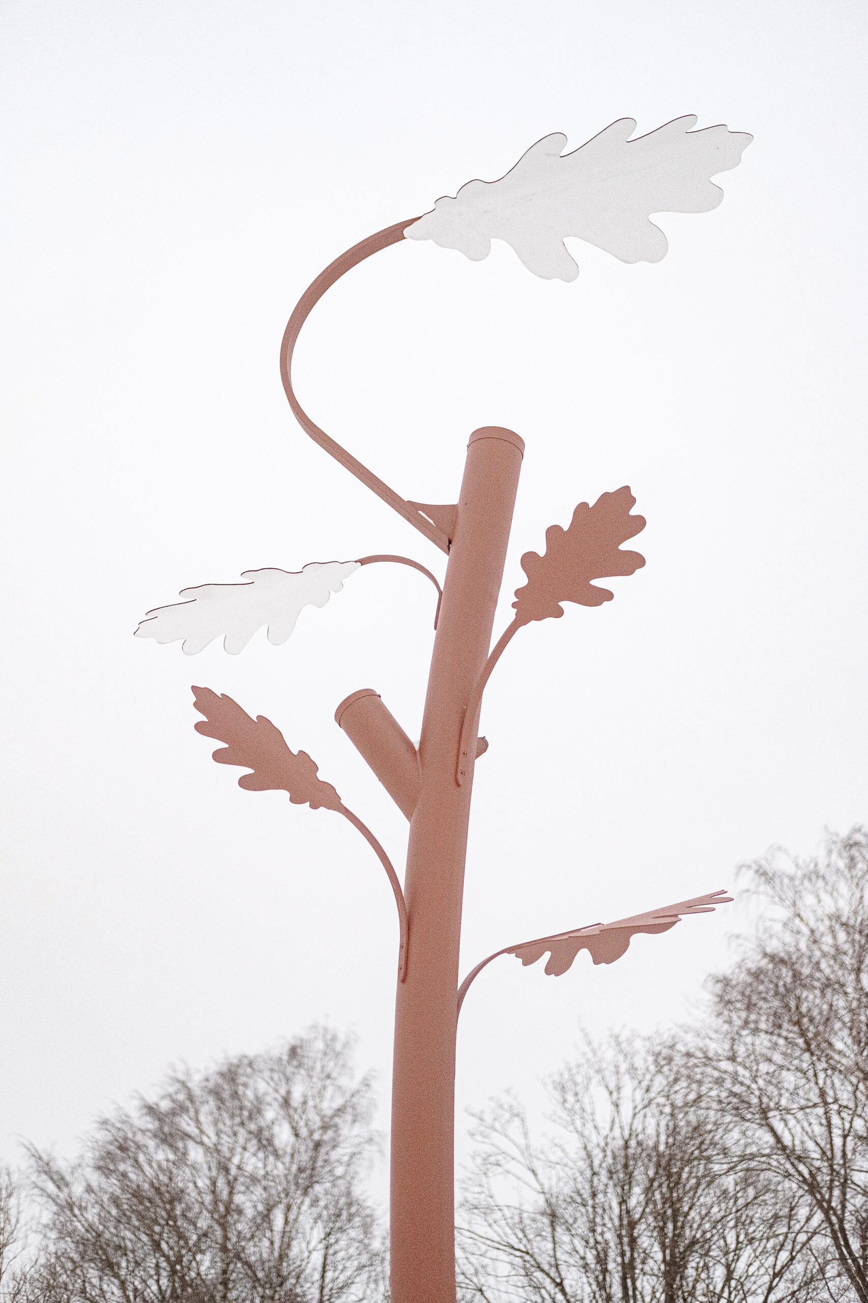 Oak tree shaped metal outdoor pole light with leaf-shaped elements against a bright, overcast sky with bare trees in the background