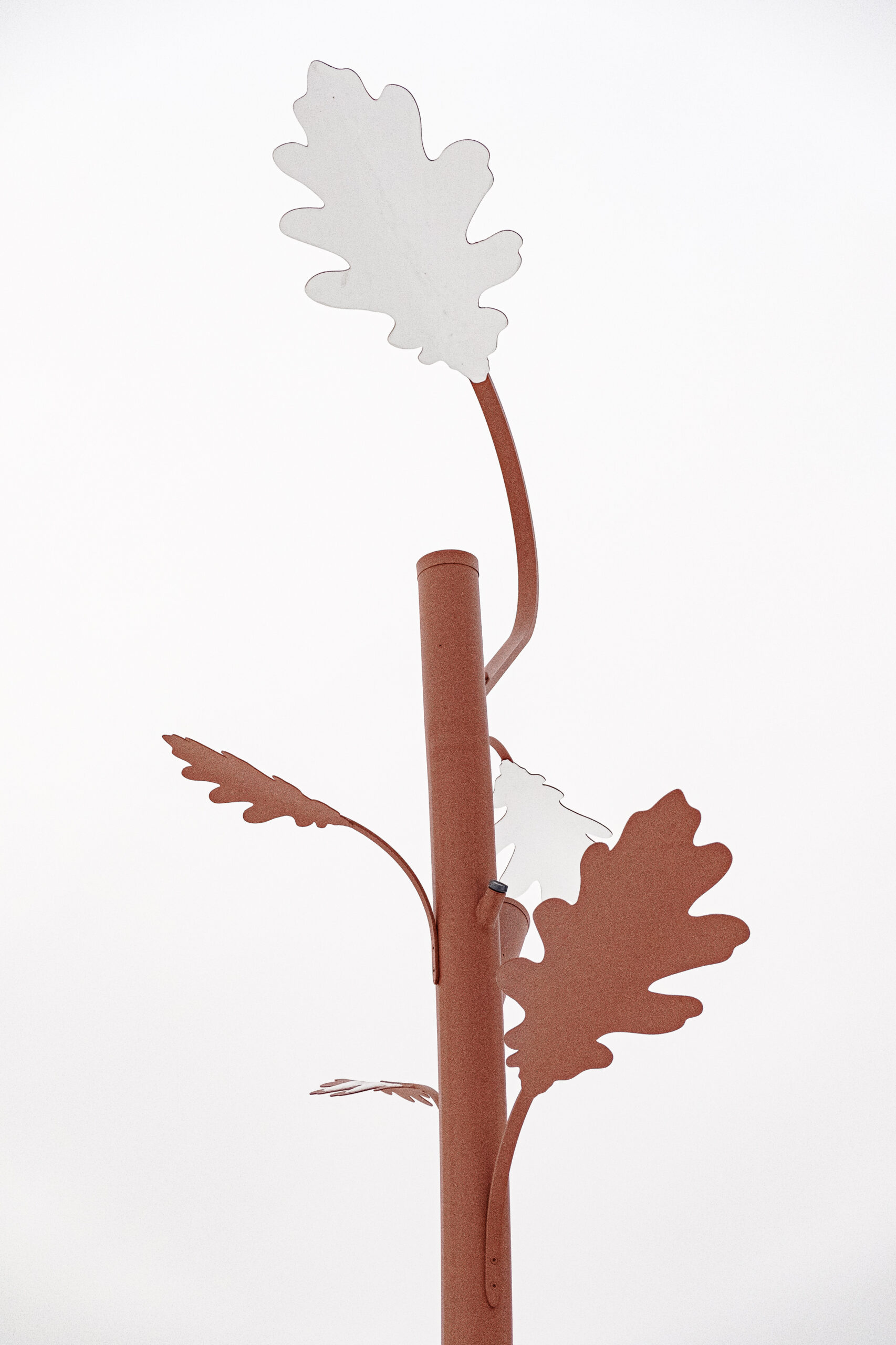 Oak tree-shaped metal custom-made outdoor light fixture in public park, with leaf-shaped elements against a bright, overcast sky with bare trees in the background