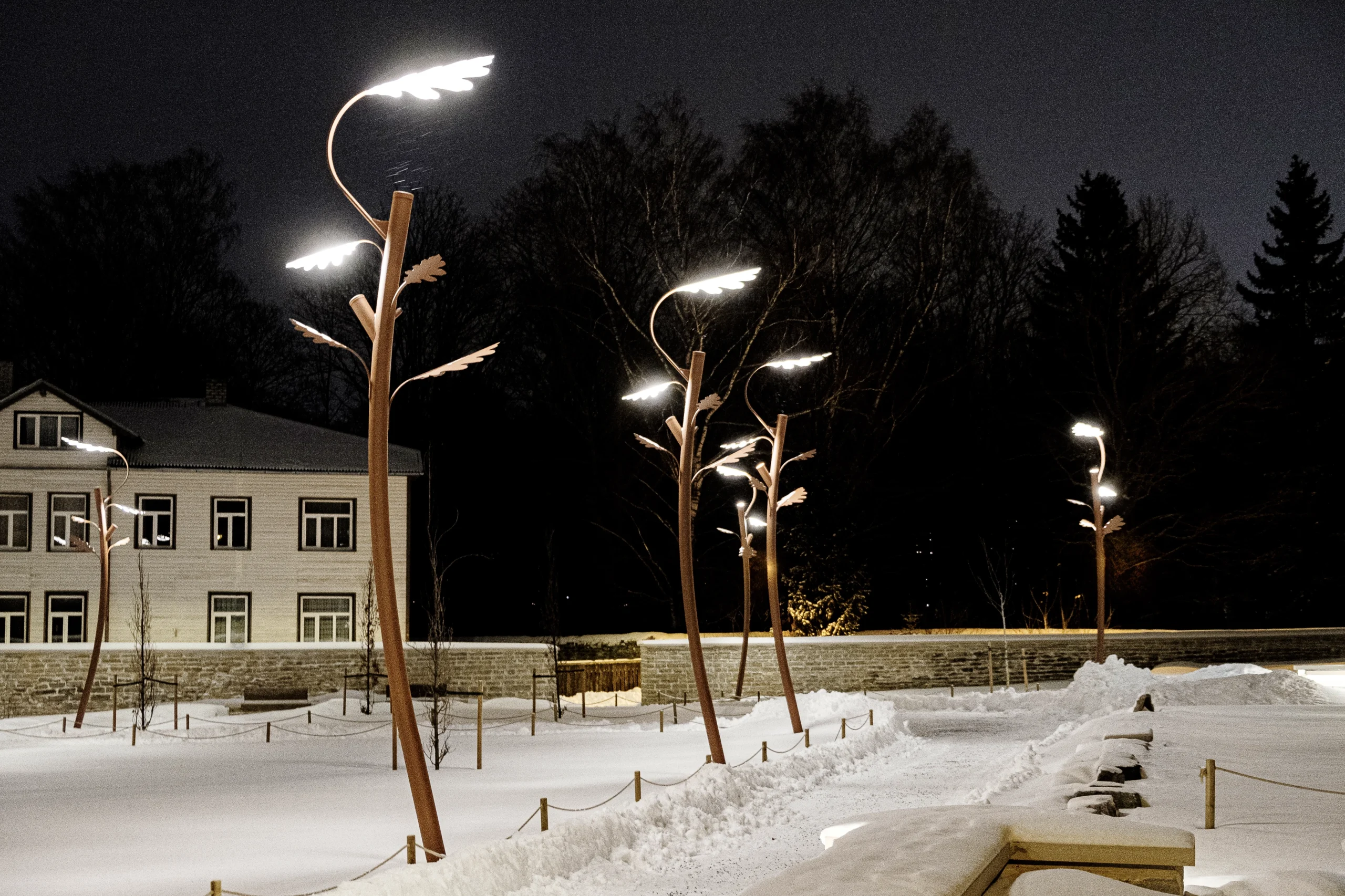 Snow-covered park at night featuring an illuminated oak tree-shaped outdoor light, stone wall, and house in the background.