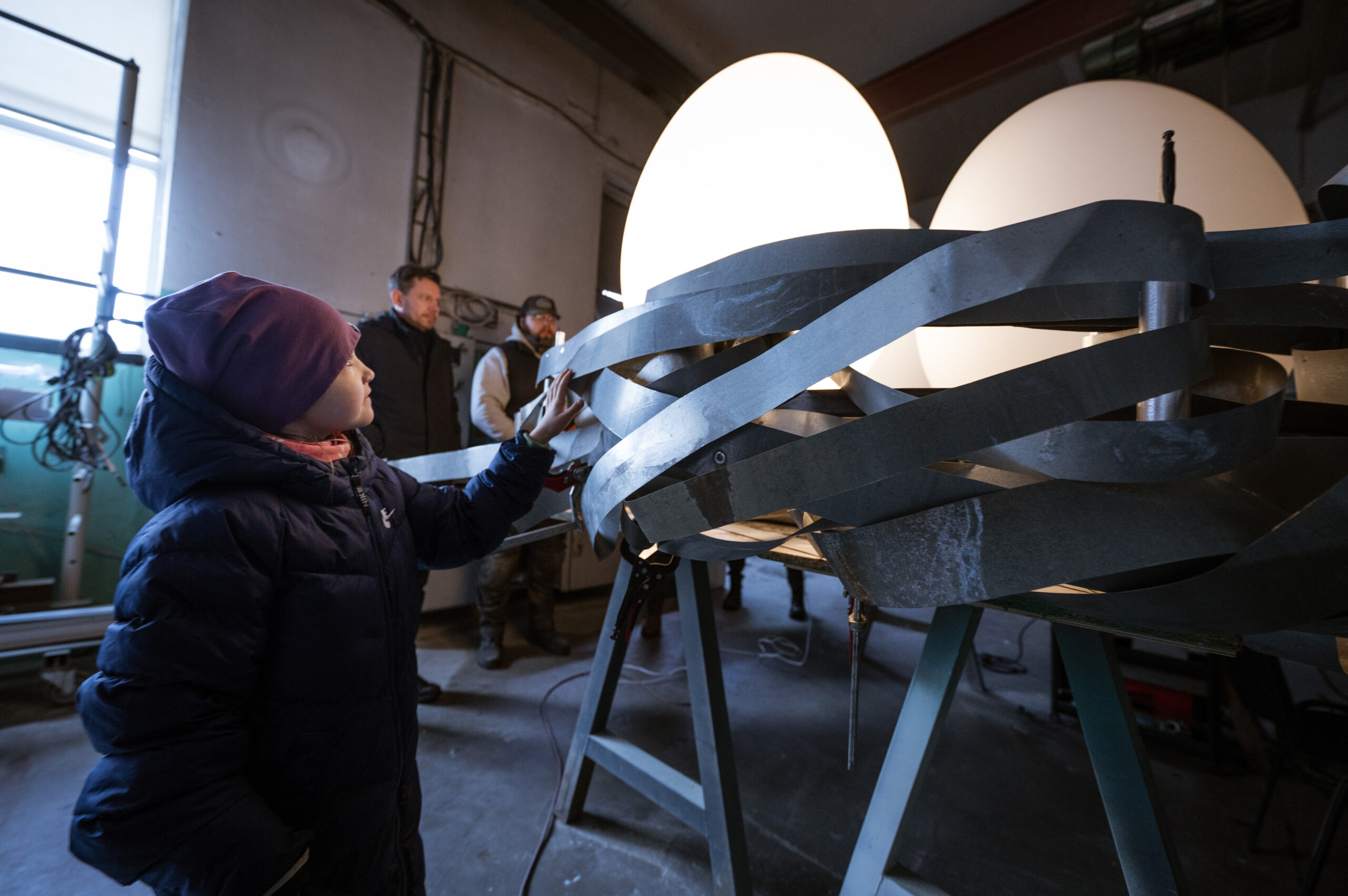 A child touches a large, illuminated nest eggs art installation indoors, with three adults observing in the background