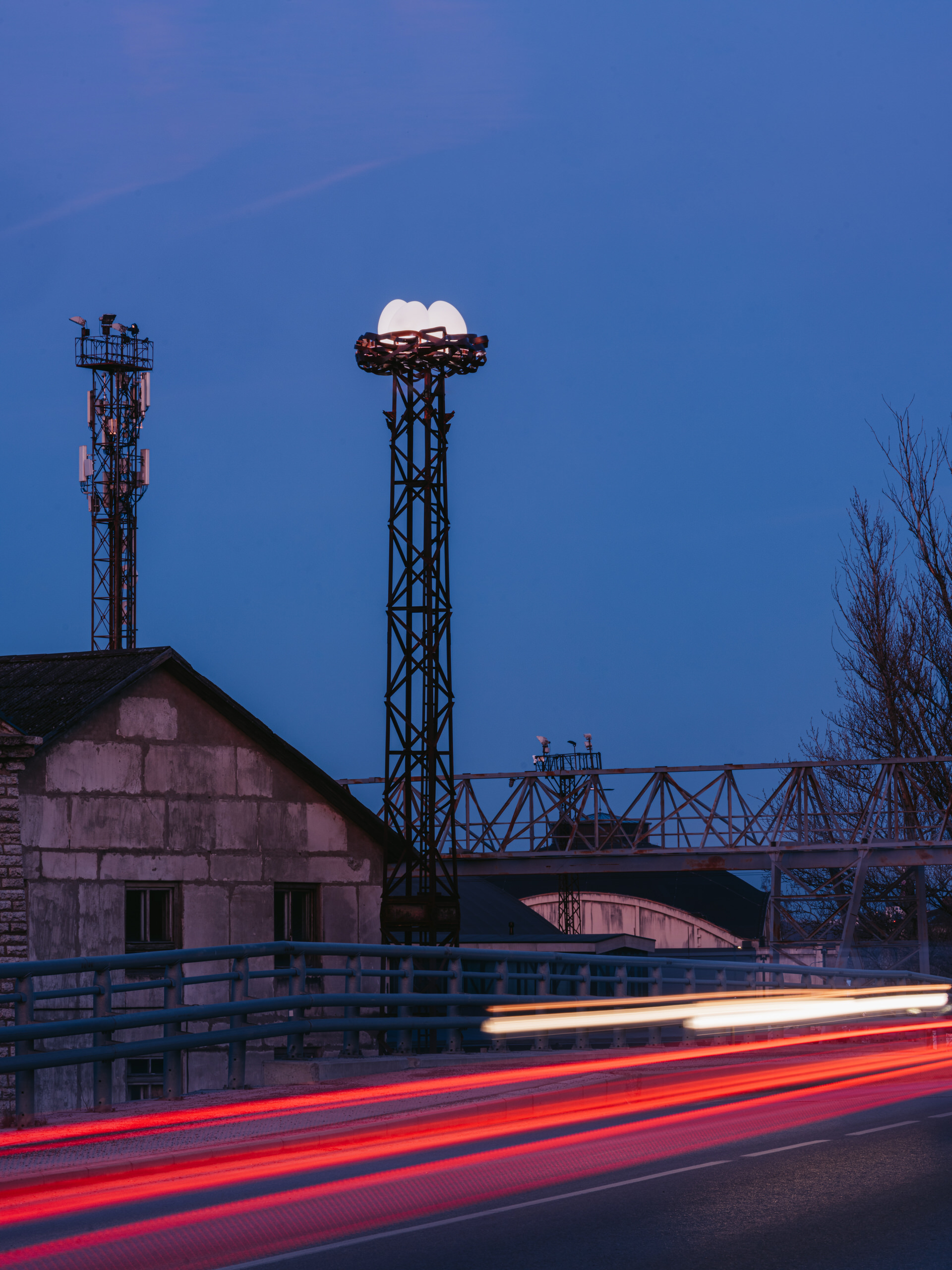 The twilight scene with a stork’s nest-inspired bespoke lighting installation in public space, showcasing three UV-resistant glowing rotomolded eggs atop a metal tower. Below, the blur of a passing car’s lights contrasts with the serene blue dusk, highlighting a historic stone building and industrial setting