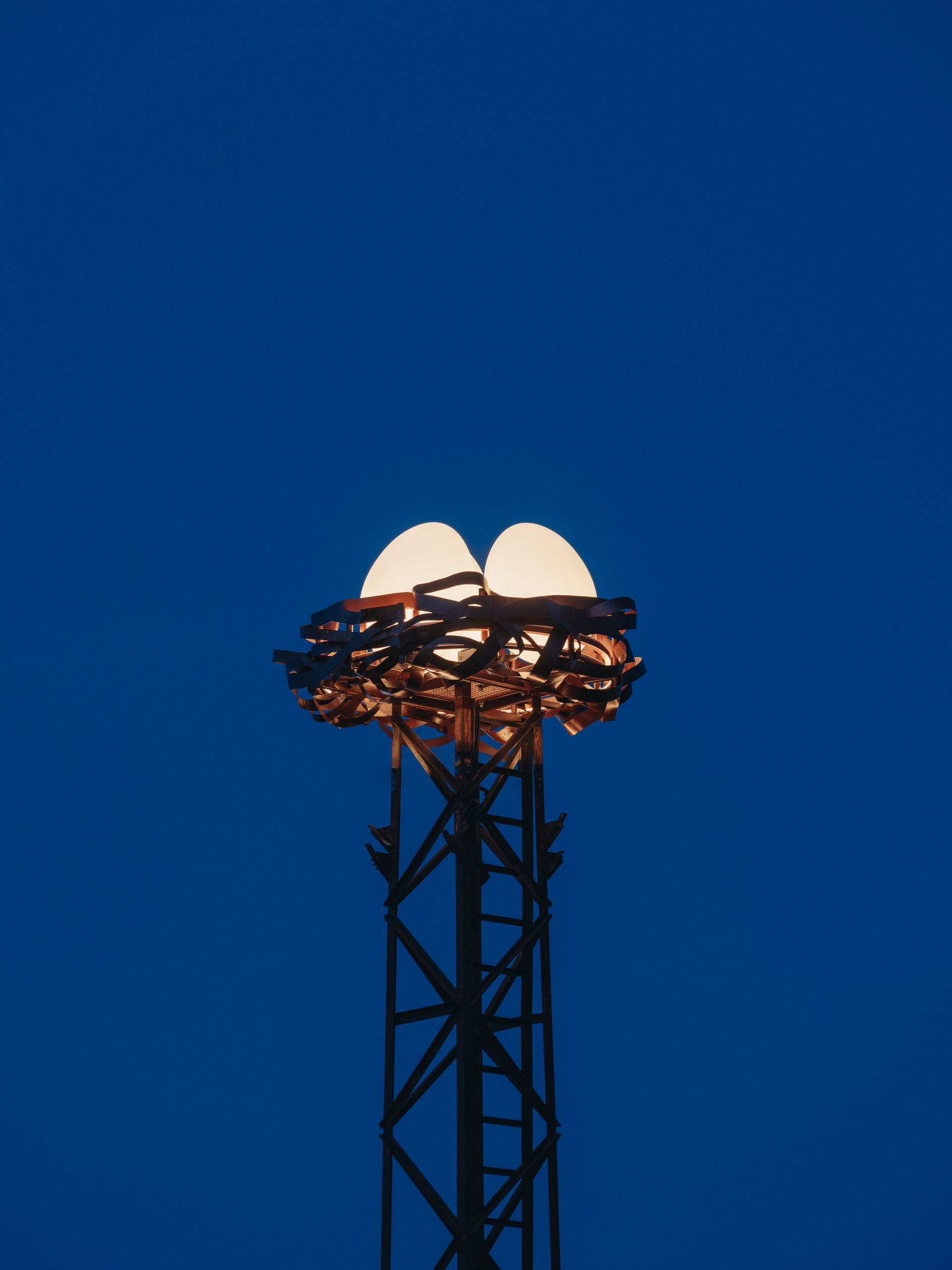 Close-up of a stork’s nest-inspired bespoke lighting installation at dusk, with three white rotomolded and UV-resistant glowing eggs in a metal weave atop a steel tower against a deep blue sky.