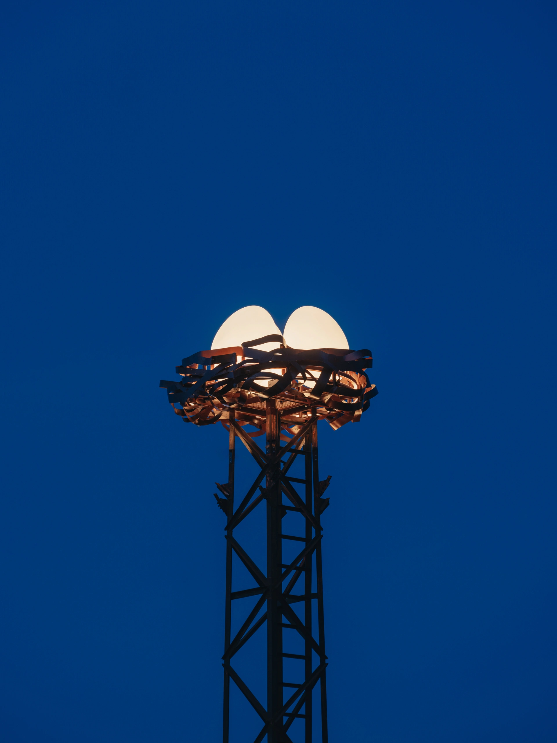 Close-up of NEST urban light installation with glowing rotomolded eggs in a woven metal nest atop a steel tower against a deep blue sky.