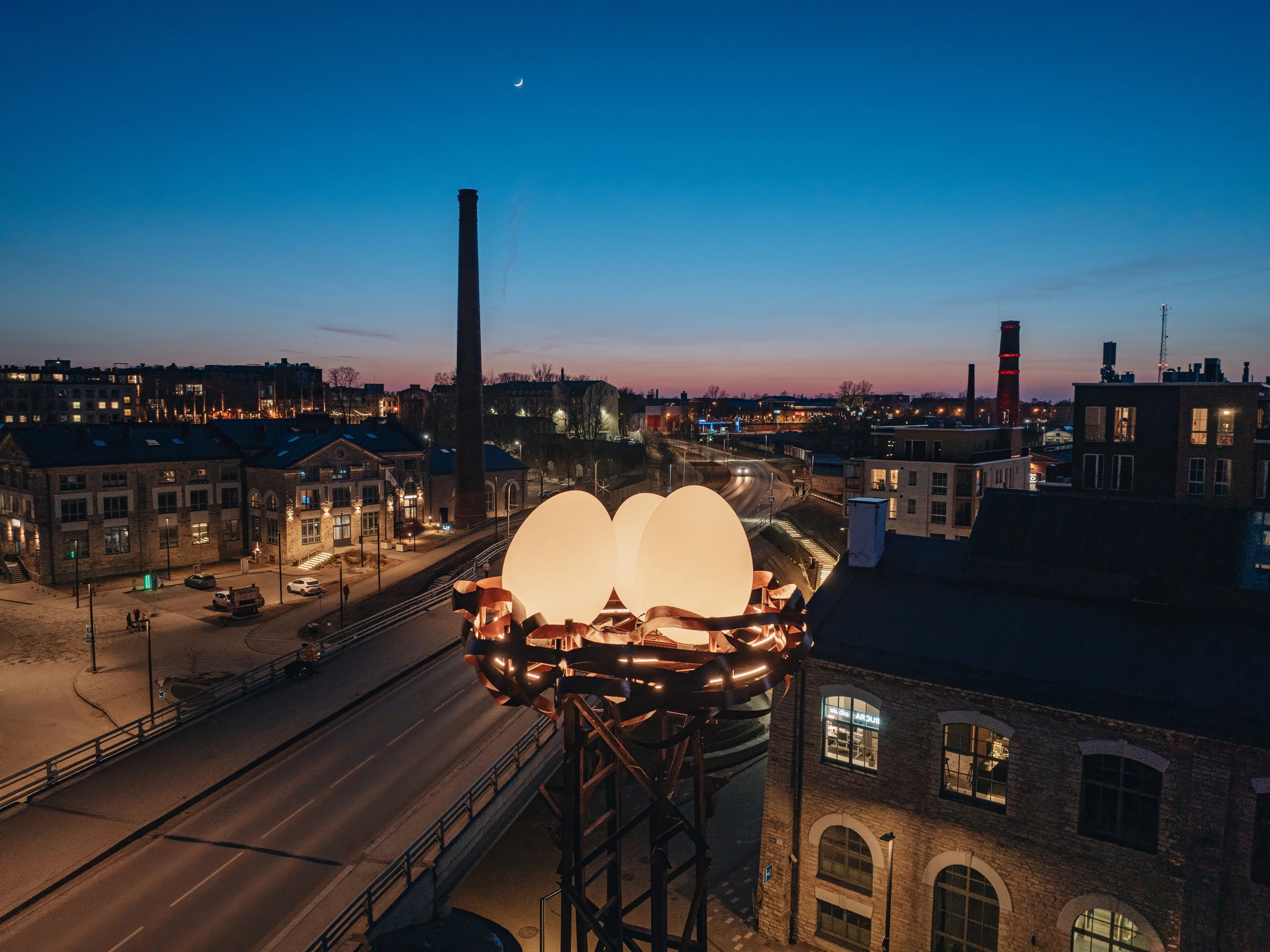 Close-up of the NEST urban light installation, featuring glowing sculptural eggs atop a steel structure, set against an industrial cityscape with high chimneys and a colorful evening sky