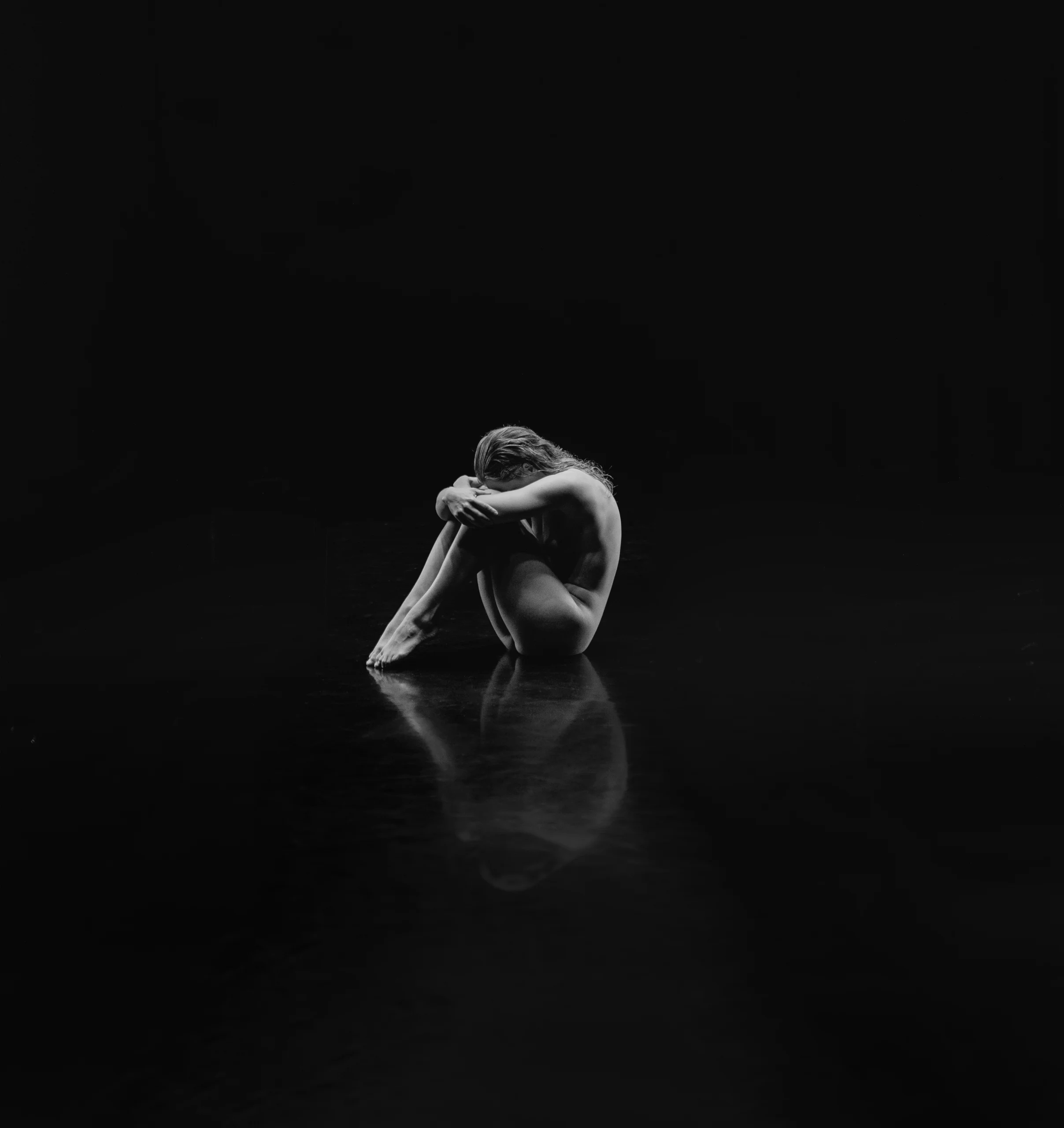 Monochrome image of a woman in a black room, her reflection visible on the glossy floor beneath HEXA lighting.