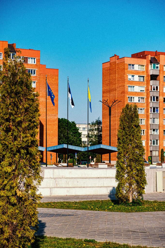 Ahtme's rebuilt central square framed by conifers, with Estonian, EU and Ukrainian flags and a Cornflower pole