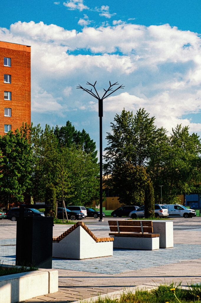A Cornflower pole rises over the square's benches and bins under cloud, part of Ahtme's public square lighting