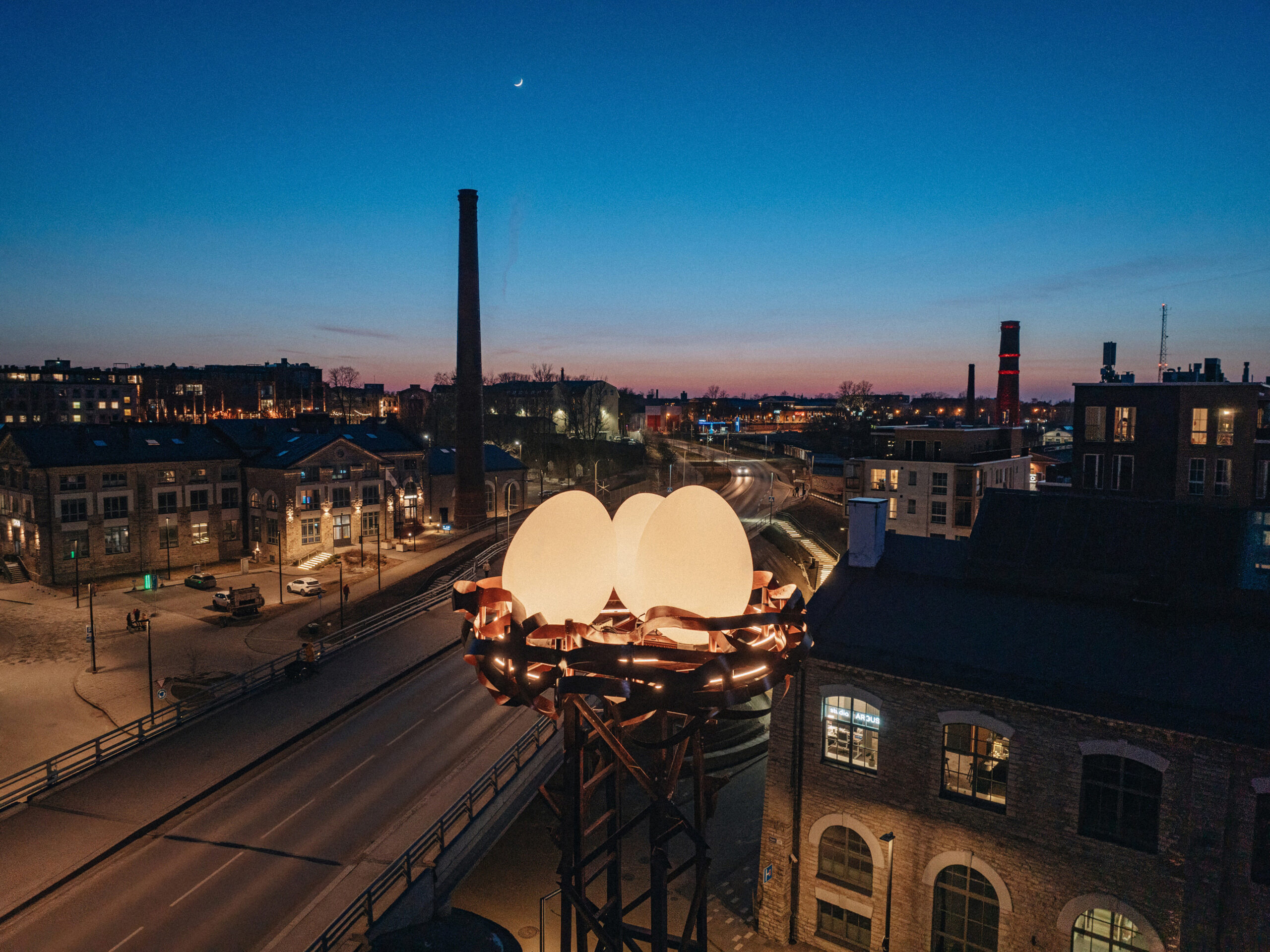 Nest with glowing eggs on a steel tower above the Noblessner rooftops at twilight, Tallinn