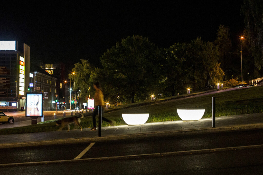 Timpan illuminated street furniture glowing at night as a person walks a dog, Tartu