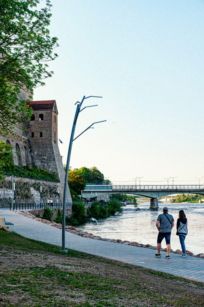 People walking the Narva riverside promenade past a Tree luminaire, castle and bridge beyond