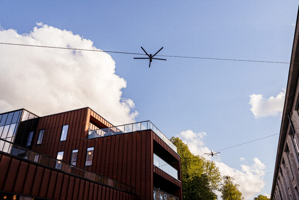 Blackbird catenary lights on their wires above the Corten-clad building and its glass roof terrace, white clouds behind