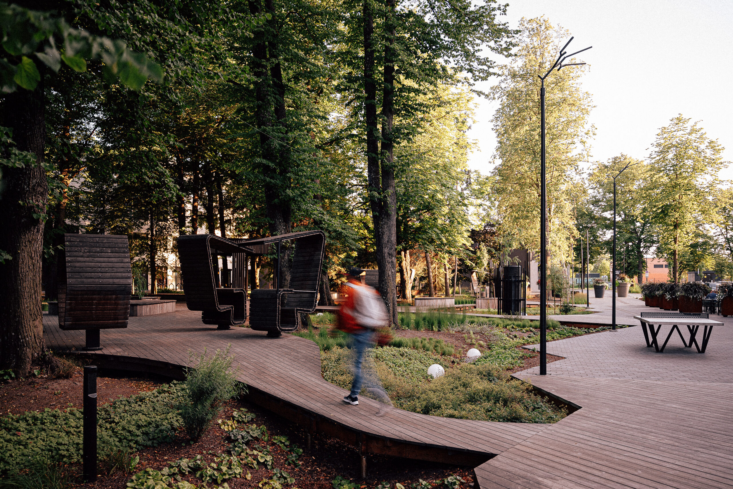 Branch and Buoy park lighting in Tallinn, a walker in an orange jacket crossing the boardwalk past high-backed timber seats