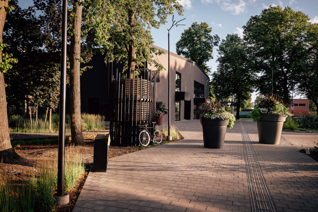 Branch luminaire over a paved path on a summer evening, with large flower planters and a bicycle parked by a tree