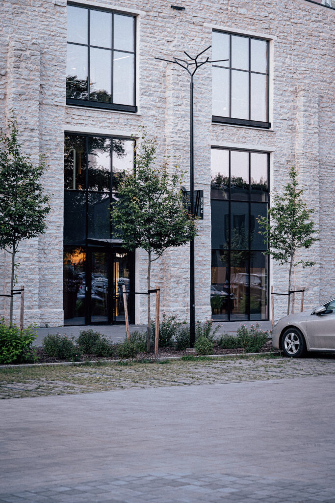 Branch luminaire between young trees at the glazed entrance of the limestone factory hall, a parked car at the kerb