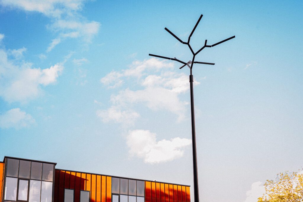 Branch urban pole-top luminaire against a bright blue sky, rising above the orange cladding of a nearby building