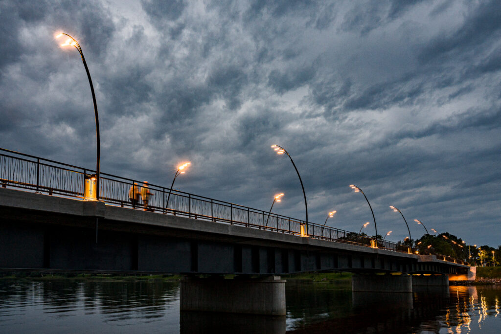 Row of lit Reed luminaires along the bridge at dusk, a pedestrian walking beneath a heavy sky.