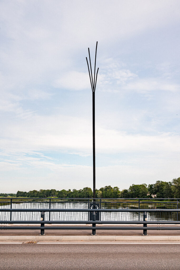 A single Reed luminaire in silhouette against a pale sky, its slender arms fanning above the bridge railing.
