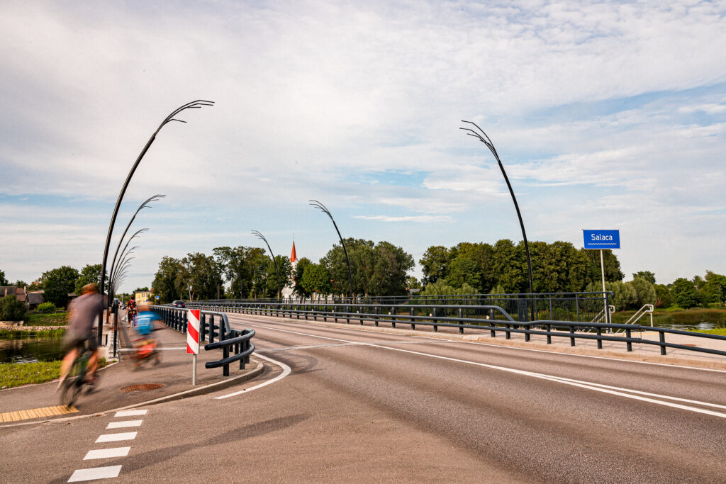 Reed pole-top luminaires lining the road bridge over the Salaca river in Salacgriva, cyclists passing below.