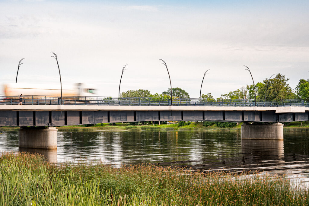 Reed lamps along the bridge railing, tall riverside grass in the foreground and a lorry passing.
